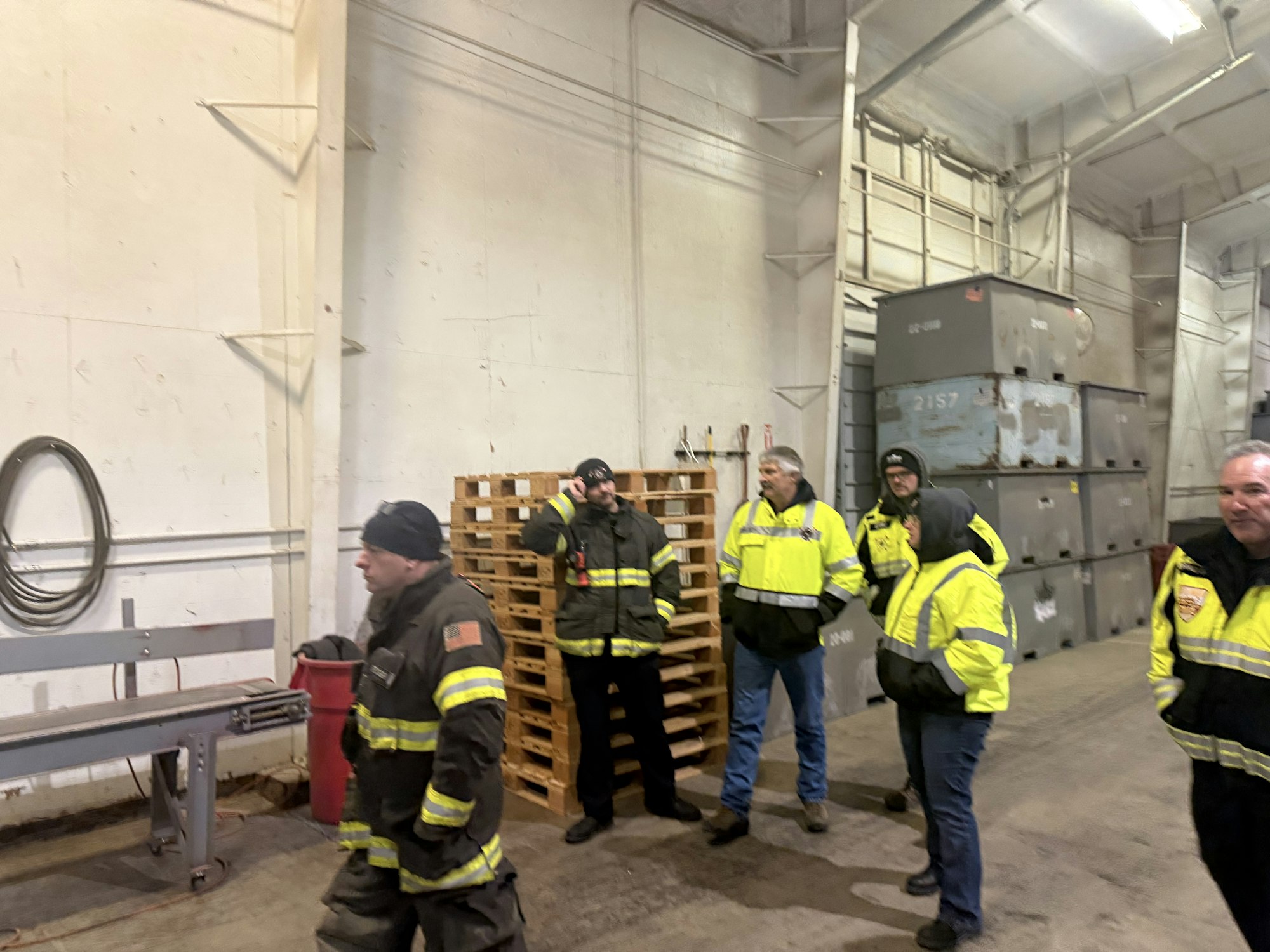 A group of individuals dressed in reflective jackets is gathered inside a warehouse, surrounded by pallets and containers during a pre-fire walk through.