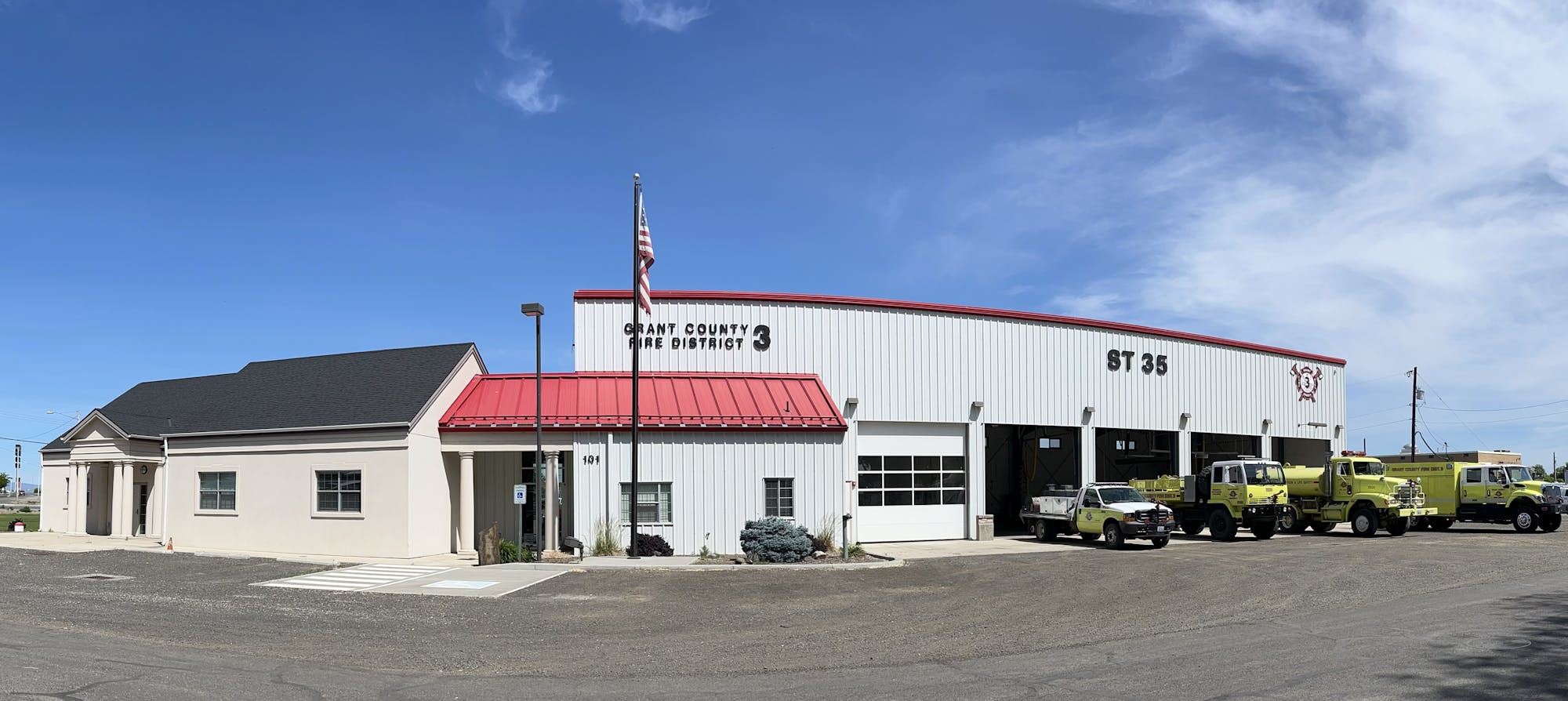 Fire station with American flag, trucks parked outside, clear sky above.