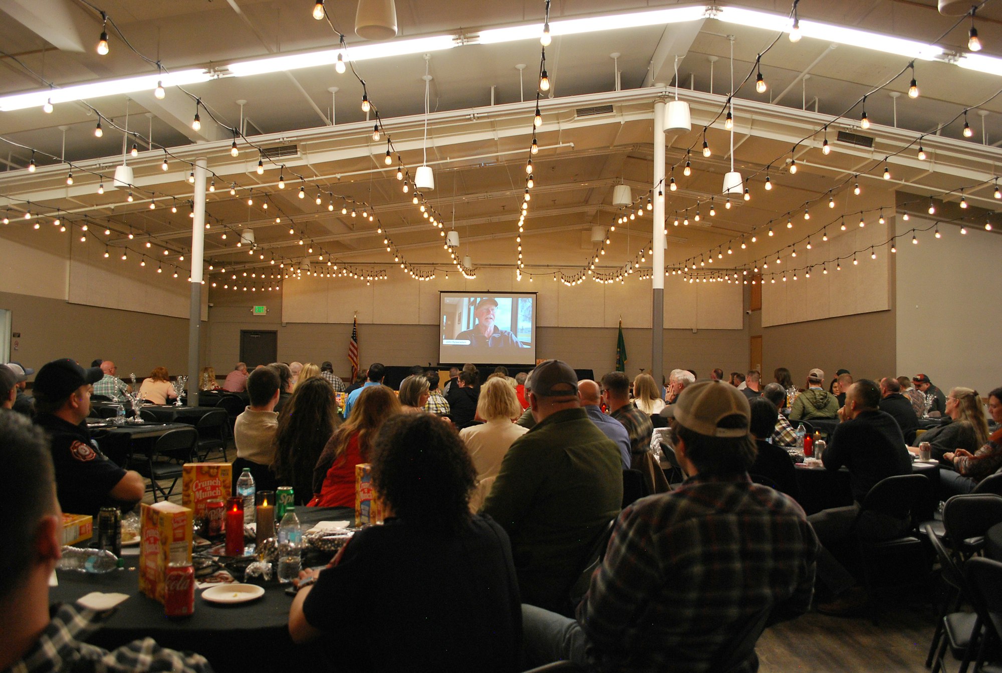 A crowded event space with attendees watching a presentation on a screen, surrounded by string lights and tables with snacks.