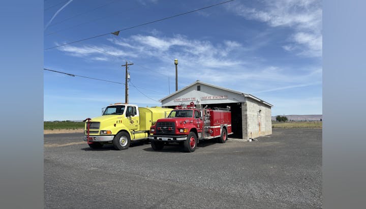 Two fire trucks parked outside a small fire station under a clear blue sky.