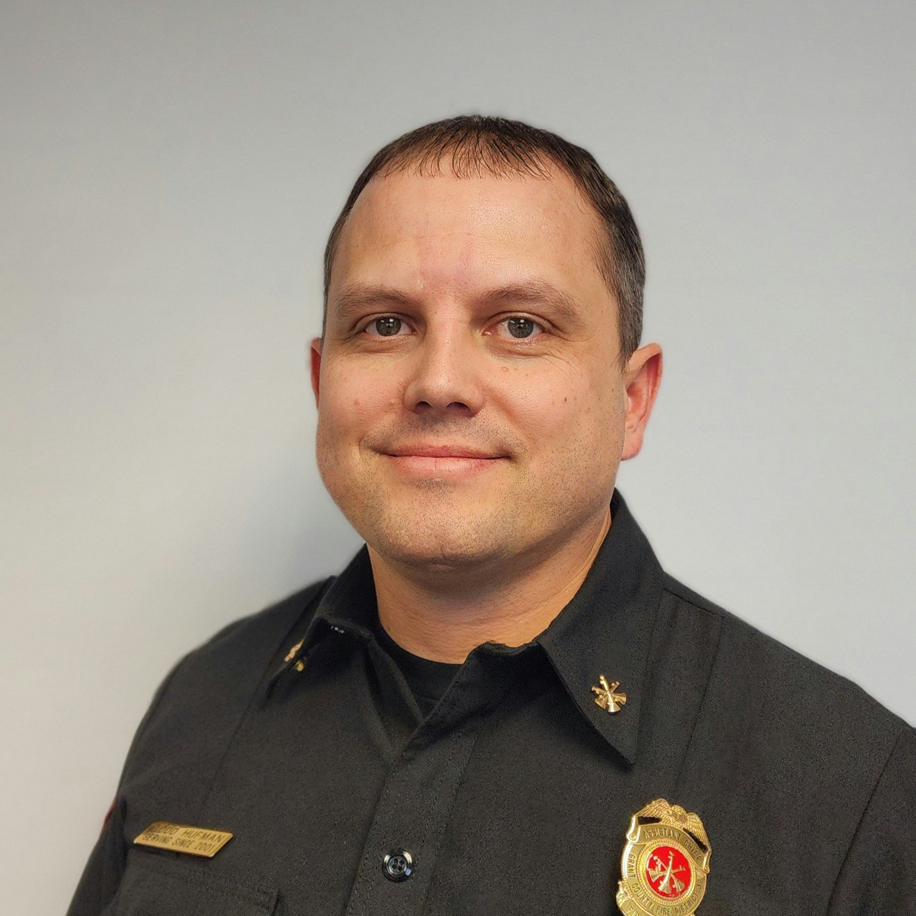 A person in a dark uniform with a badge on their chest, smiling against a plain background.