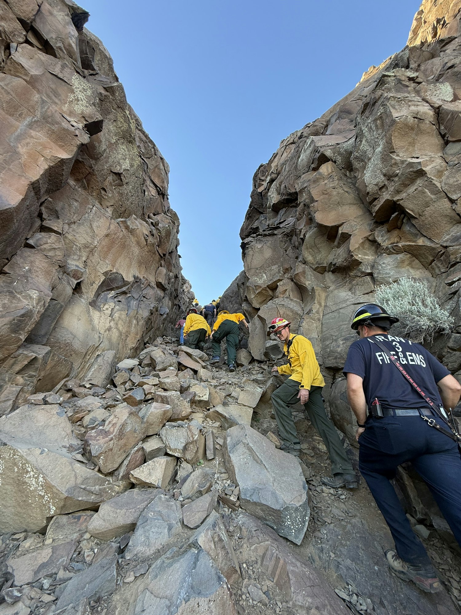 A group of people in wildland gear climbing through a rocky terrain under a clear blue sky.