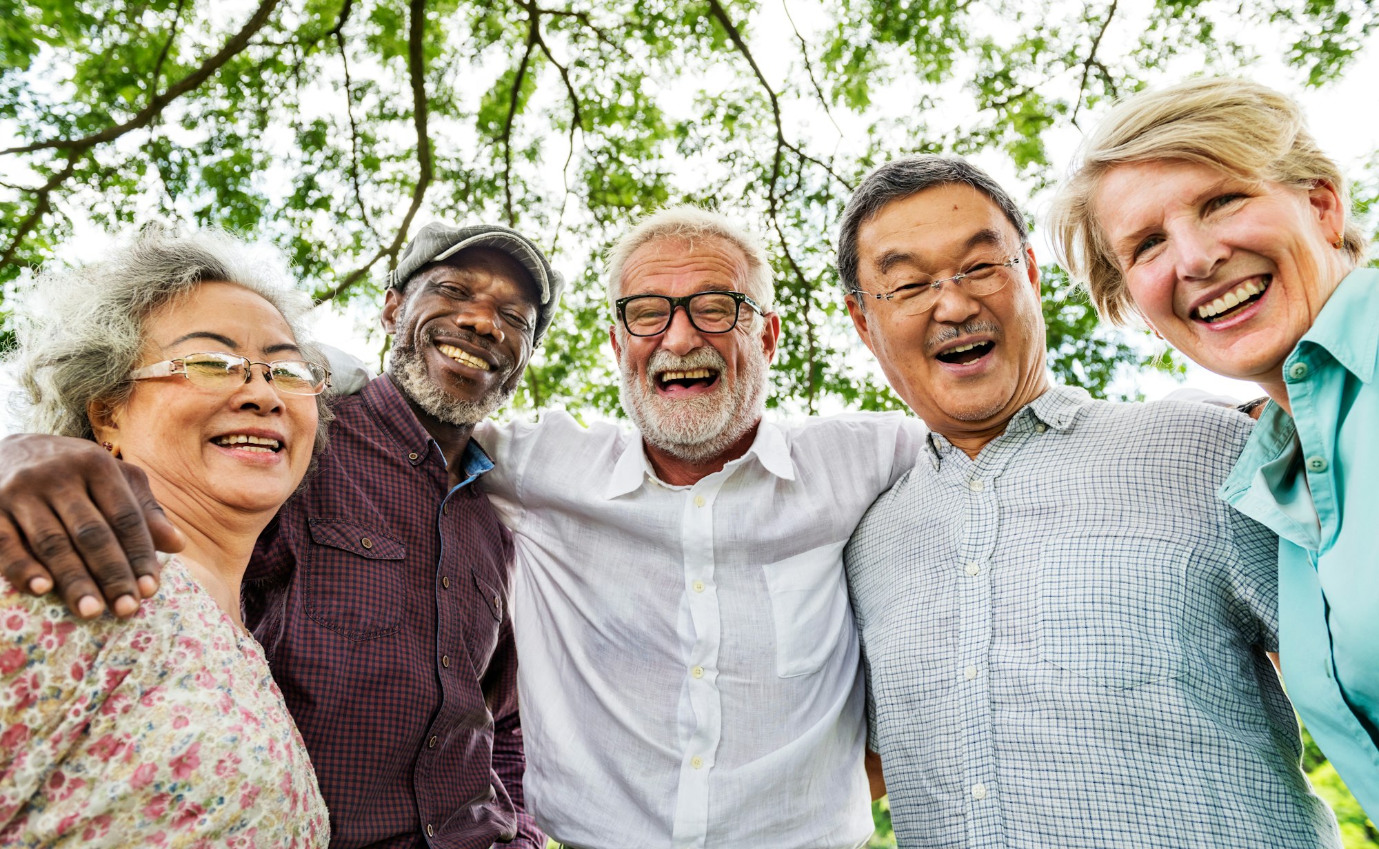 Five smiling people in a group hug outdoors, with trees in the background.