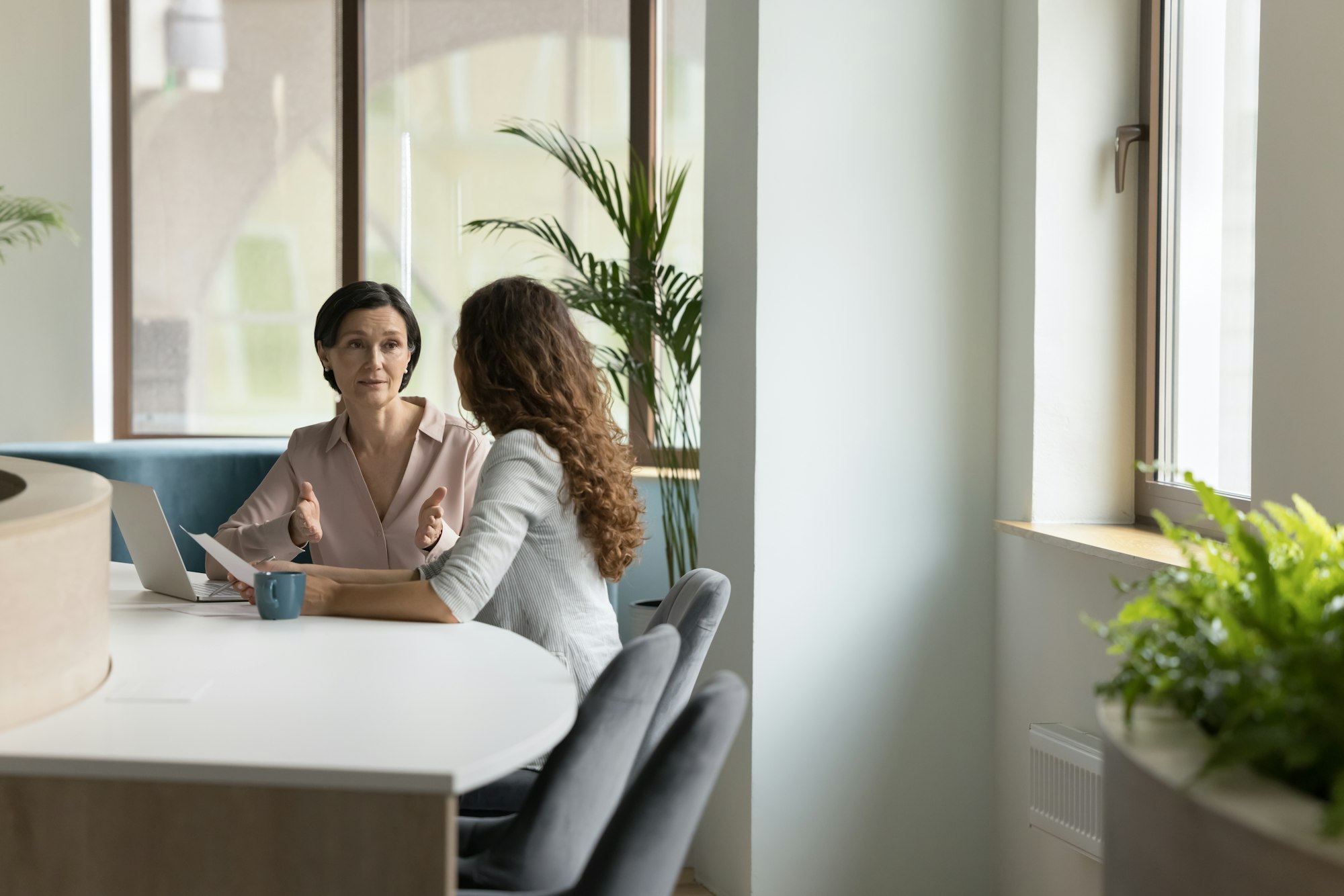 Two people are having a discussion in a bright office near a window, with a laptop and a mug on the table.