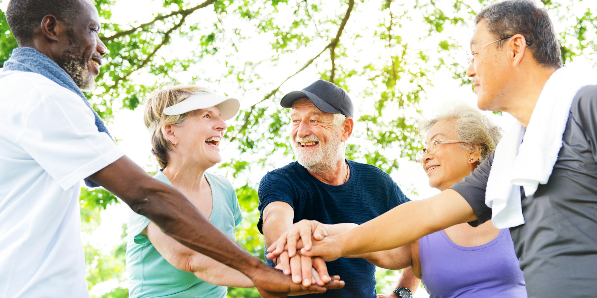 A group of smiling older adults joining hands outdoors, symbolizing friendship and team spirit.
