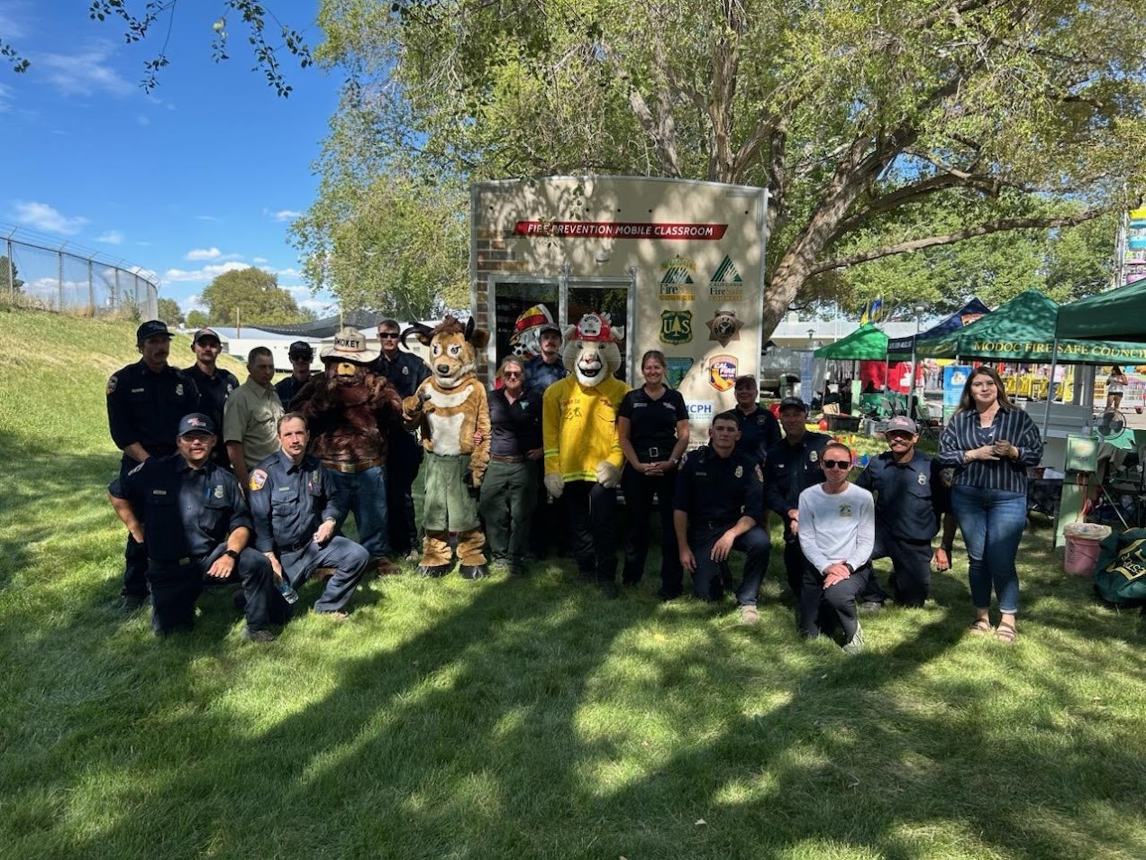 A group of firefighters, mascots, and community members gather in front of a mobile classroom at a sunny outdoor event.