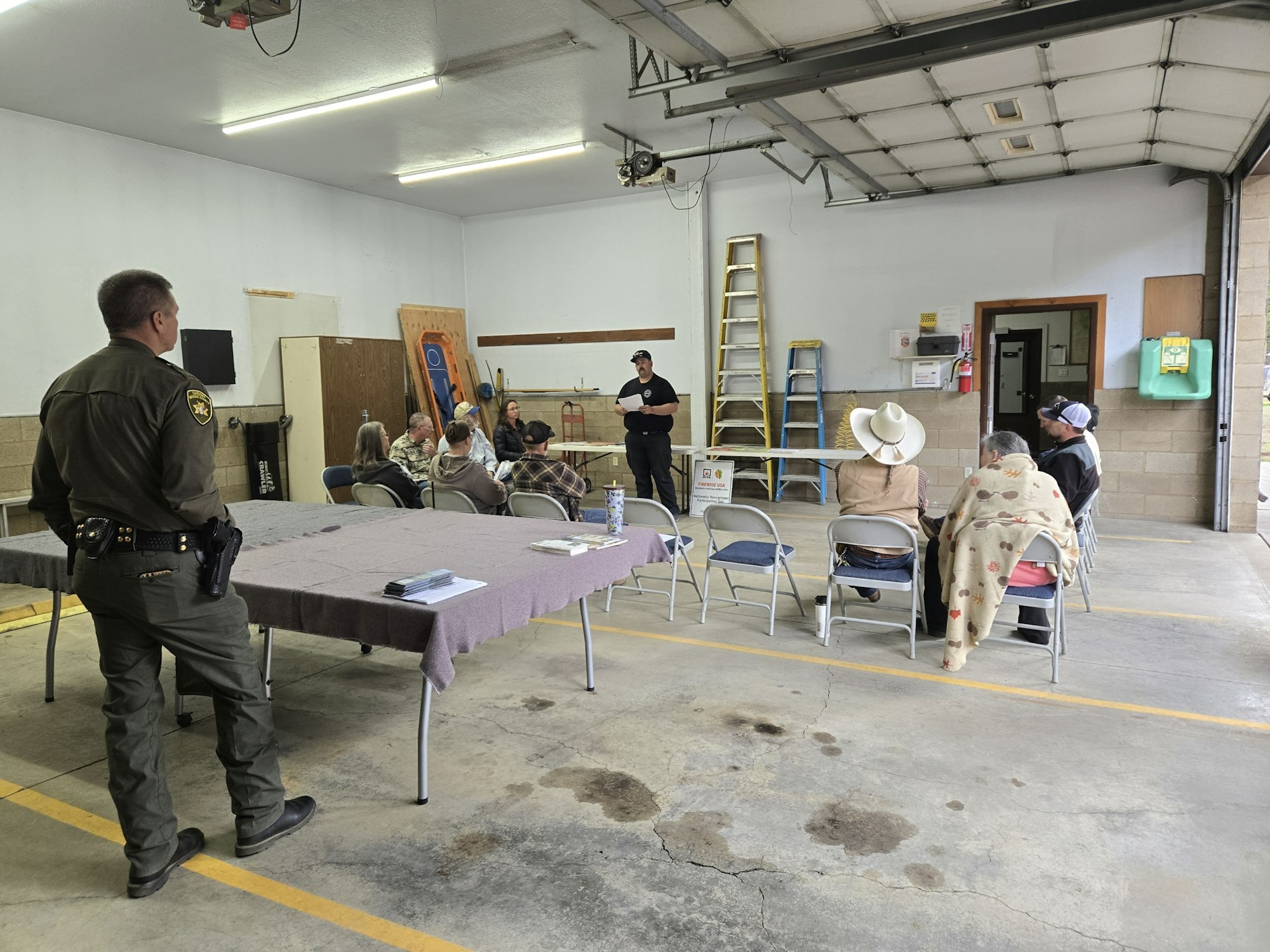 A group meets in a garage-like setting, with a uniformed officer and attendees seated around tables, listening to a speaker.
