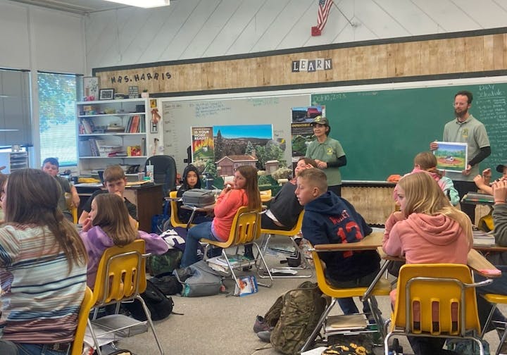 Classroom with students seated, two presenters at the front holding posters, and a "Mrs. Harris" sign above a whiteboard.