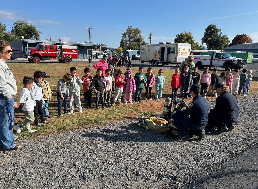 A group of children watches firefighters demonstrating equipment, with fire trucks and community members in the background.
