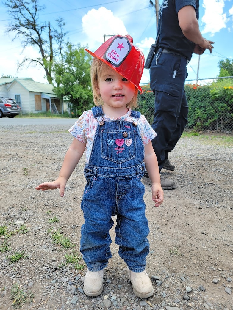 A young child wearing a fire chief hat and denim overalls stands outdoors, smiling against a sunny backdrop.