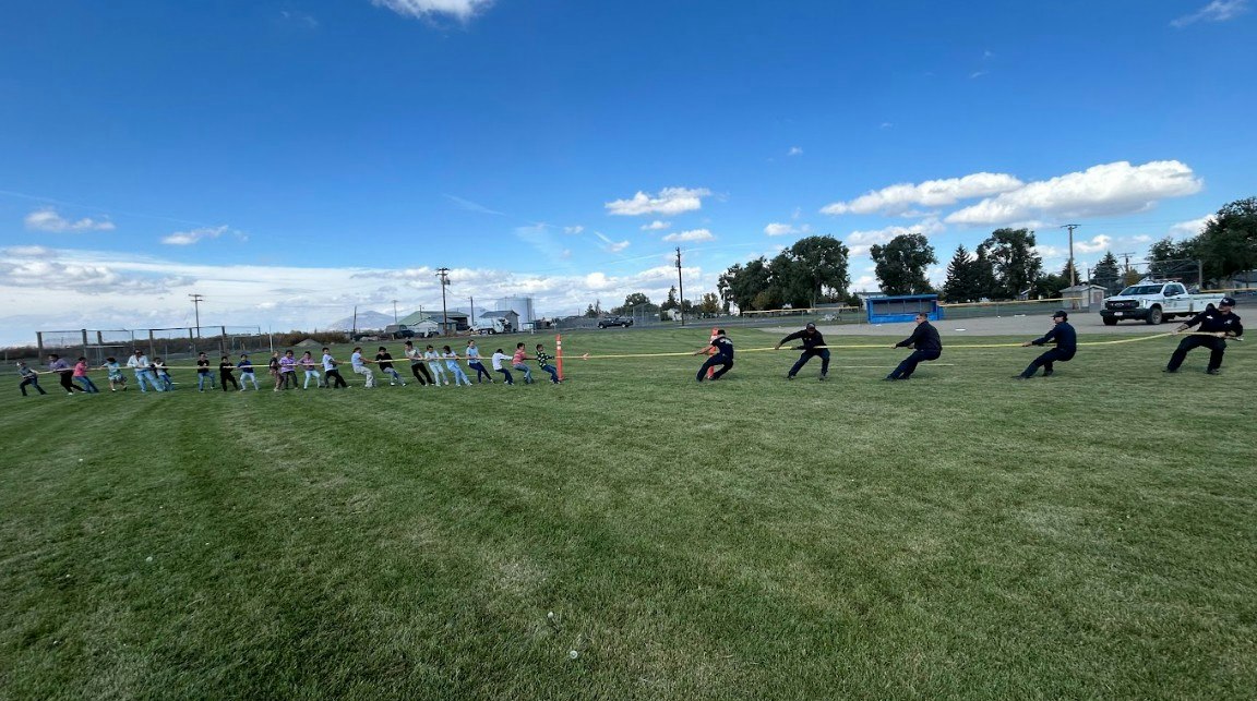 A tug-of-war competition between a group of children and adults taking place on a grassy field under a blue sky.