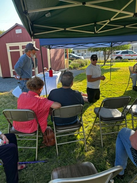 A group of people sitting under a tent, with one person speaking while others listen. Outdoor setting with grass and nearby structures.