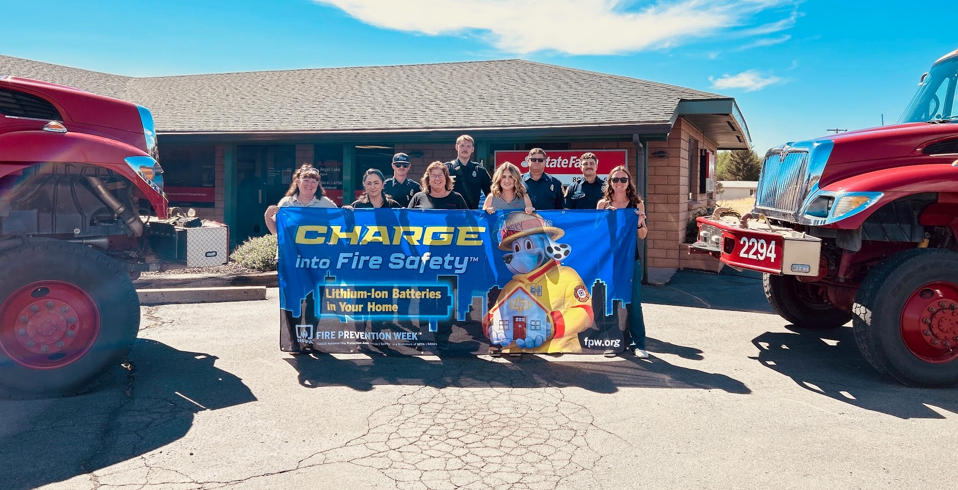 A group promotes fire safety awareness about lithium-ion batteries, holding a banner in front of fire trucks and a building.