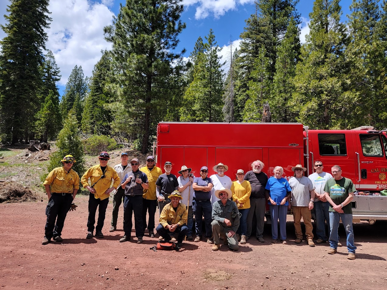 A group of people, including firefighters, pose together in front of a red fire truck in a forested area.