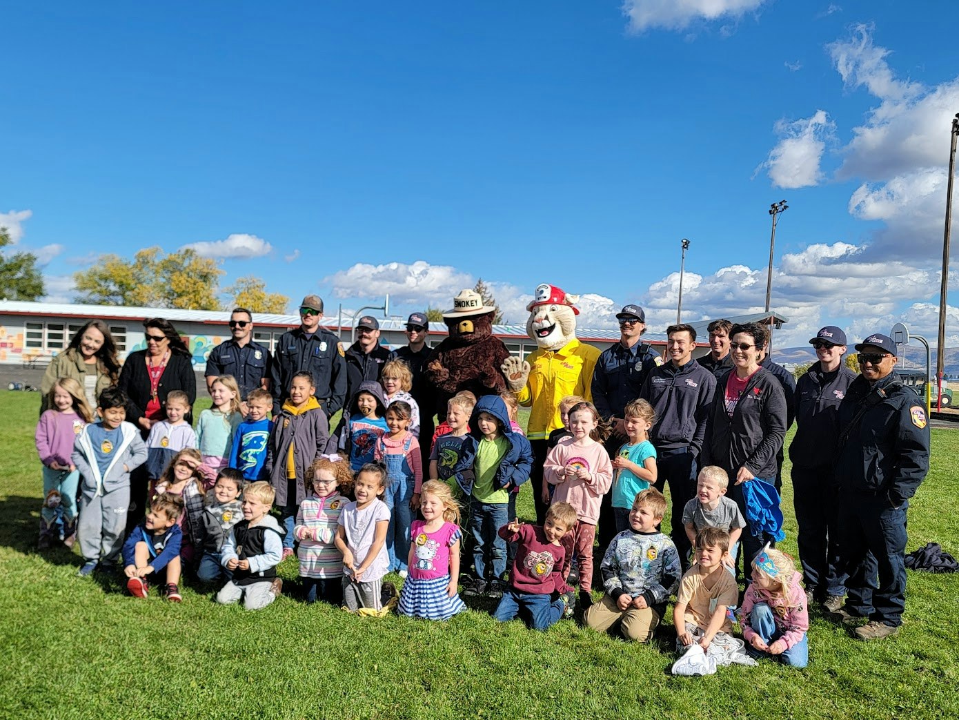 A group photo featuring children, firefighters, police officers, and mascots outdoors on a sunny day.