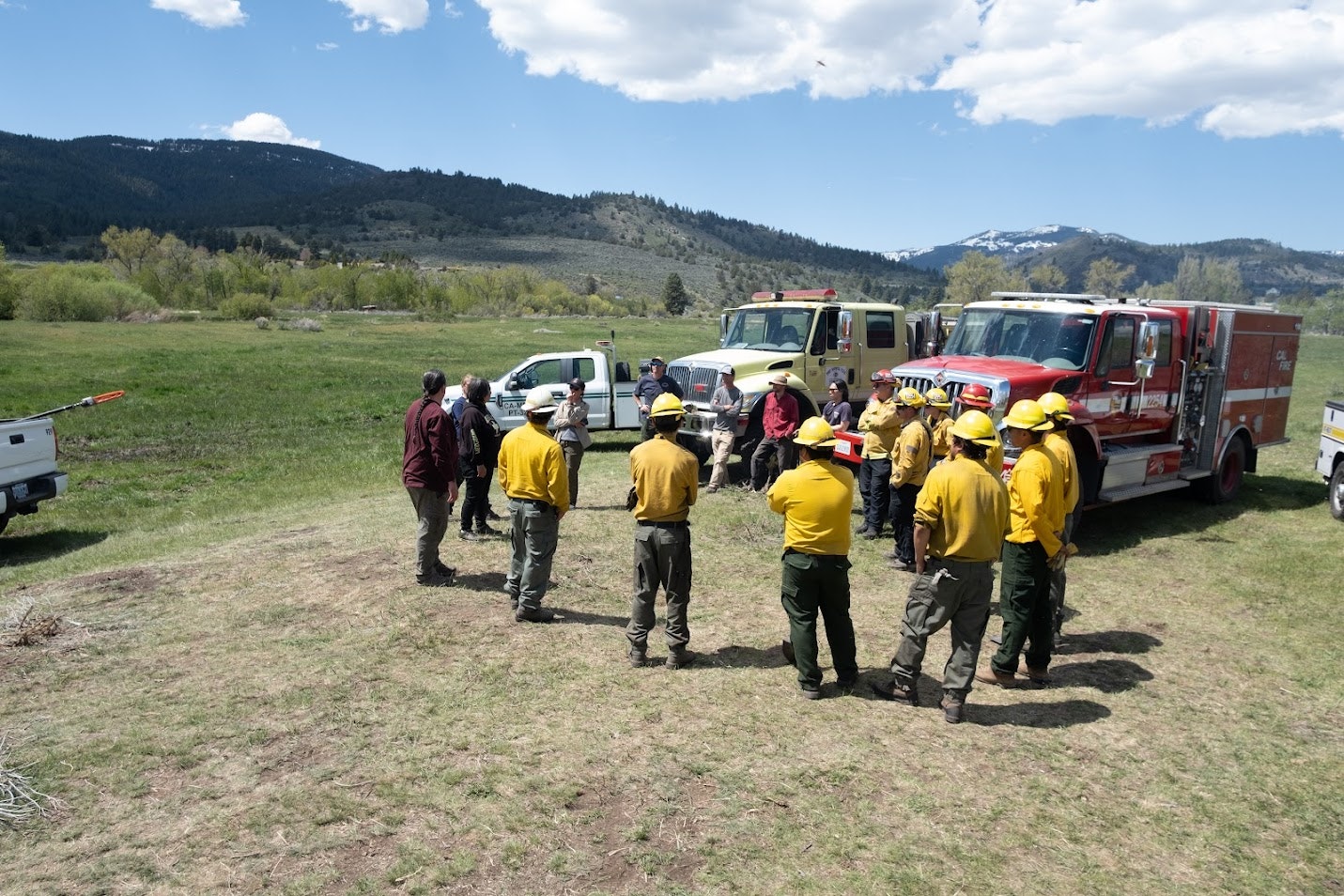 A group of firefighters and officials gather outdoors near fire trucks for a briefing or training in a mountainous area.