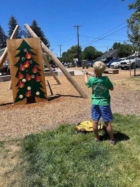 A child plays in a park, aiming at a wooden tree target decorated with colored ornaments on a sunny day.