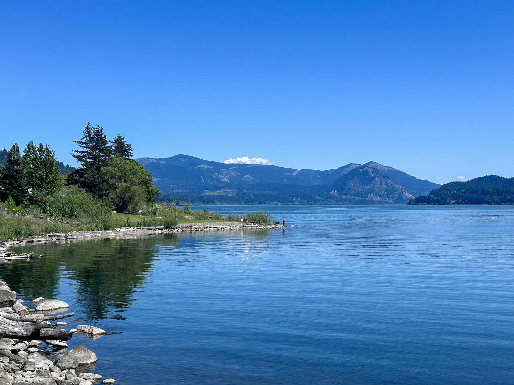 A serene lake view with mountains in the background, blue skies, and calm waters reflecting greenery and a person fishing.