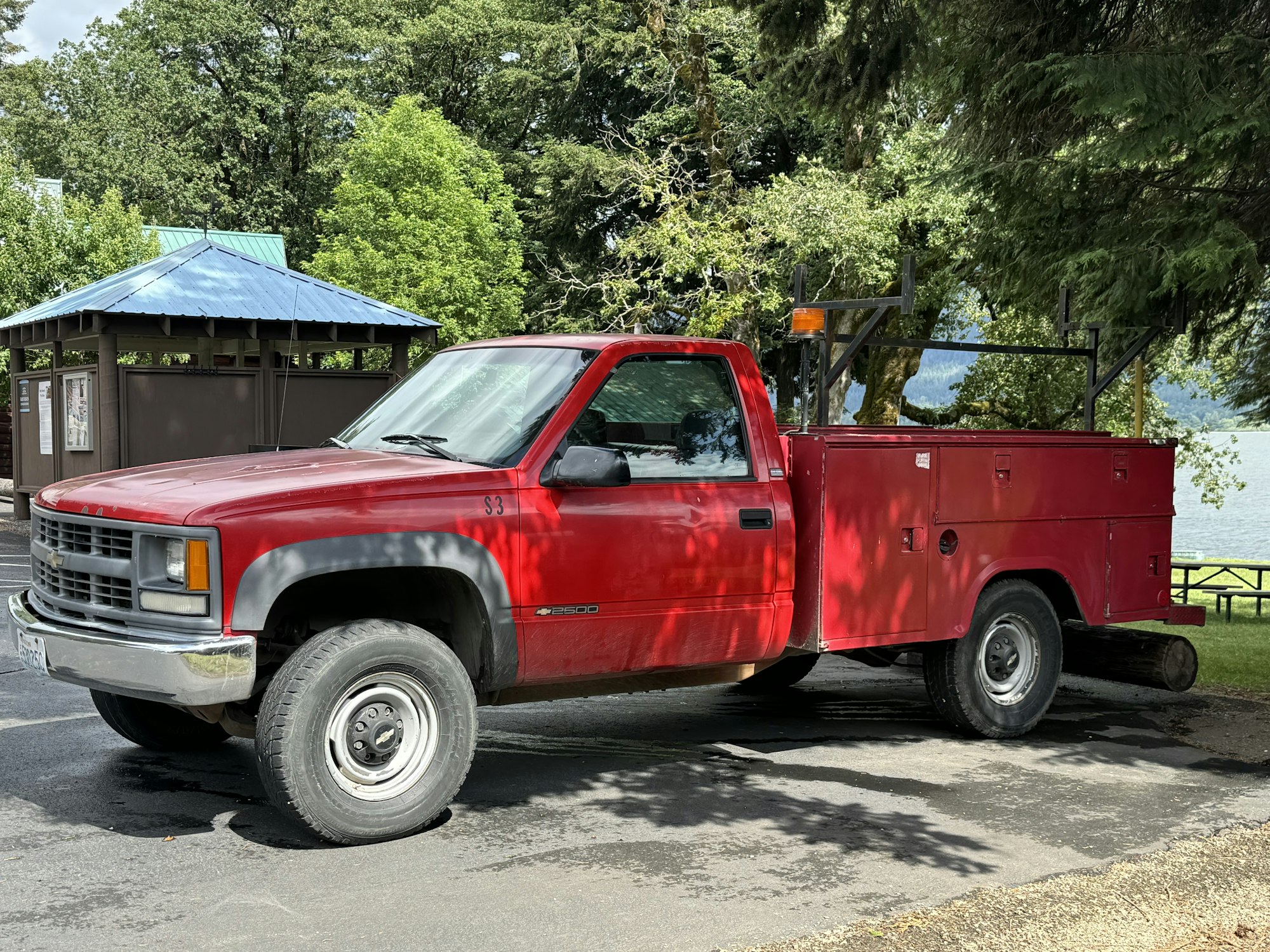 A red utility pickup truck parked near trees and a gazebo, possibly by a lake.