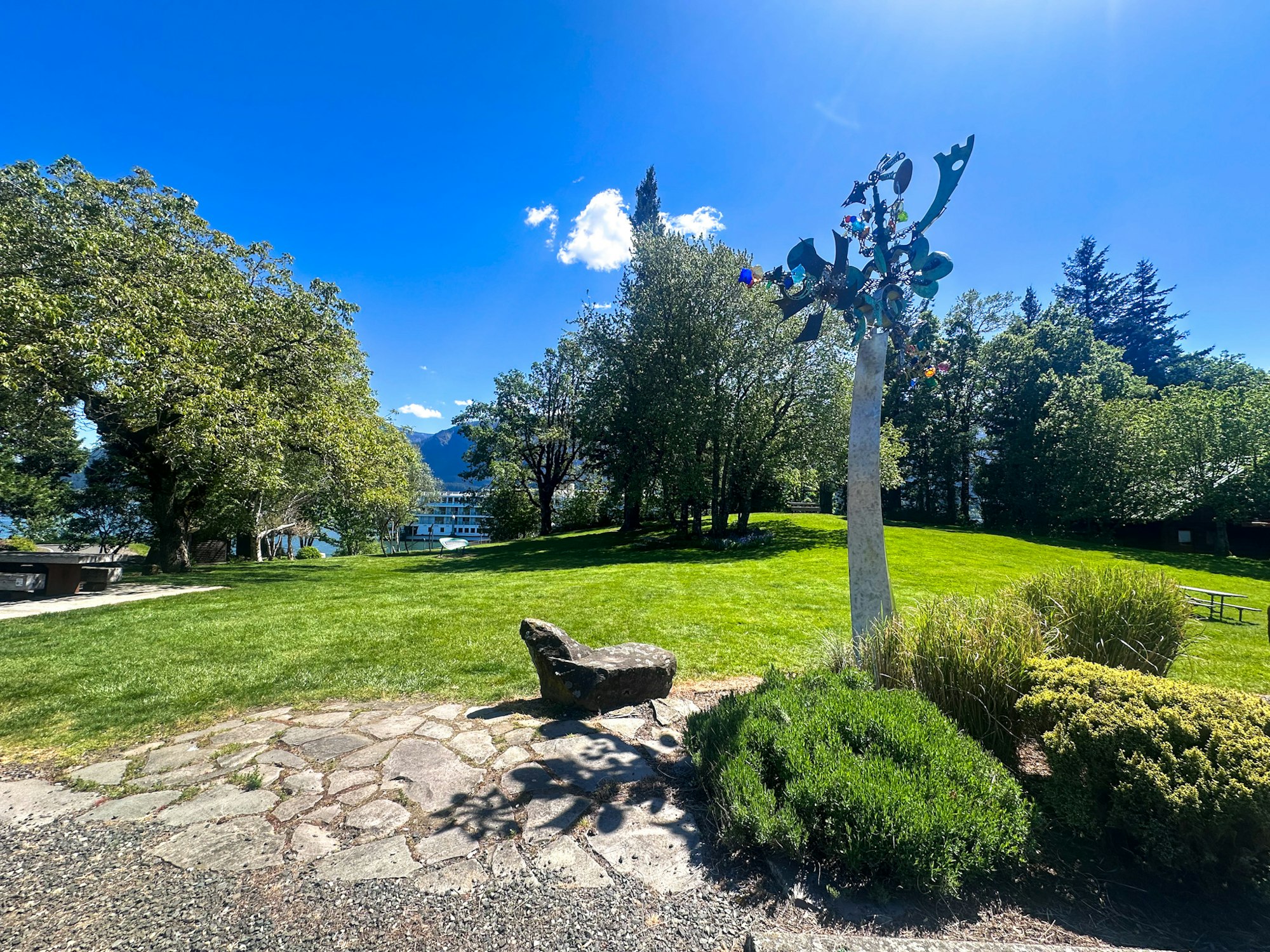 A sunny park scene features green grass, trees, stone pathways, and a colorful sculpture against a blue sky.