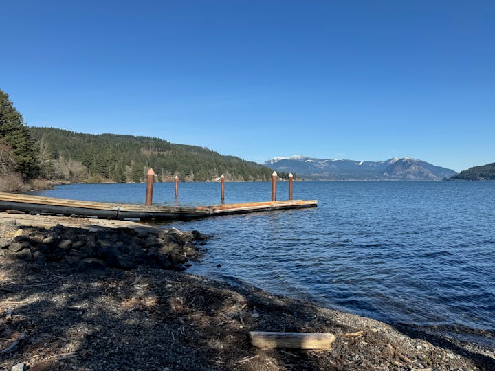 A serene lakeside with a jetty, surrounded by forested hills and snow-capped mountains under a blue sky.