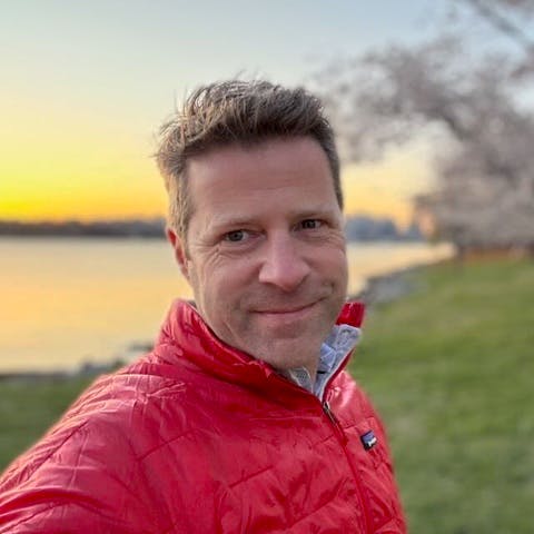 A man in a red jacket smiles for a selfie by a river at sunset, with cherry blossom trees in the background.