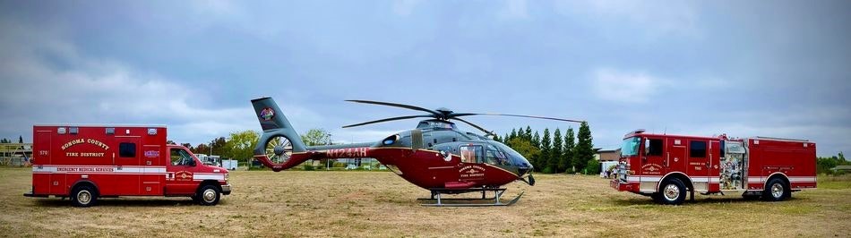 A red ambulance, rescue helicopter, and fire truck on a grassy field.