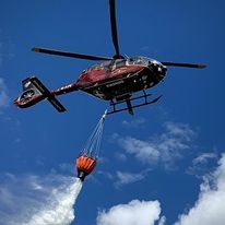 A helicopter drops water from a bucket against a blue sky.