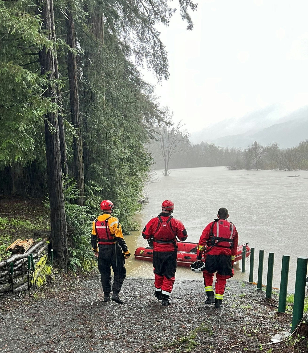 Three people in rescue gear walk towards a flooded area with an inflatable boat, surrounded by trees.