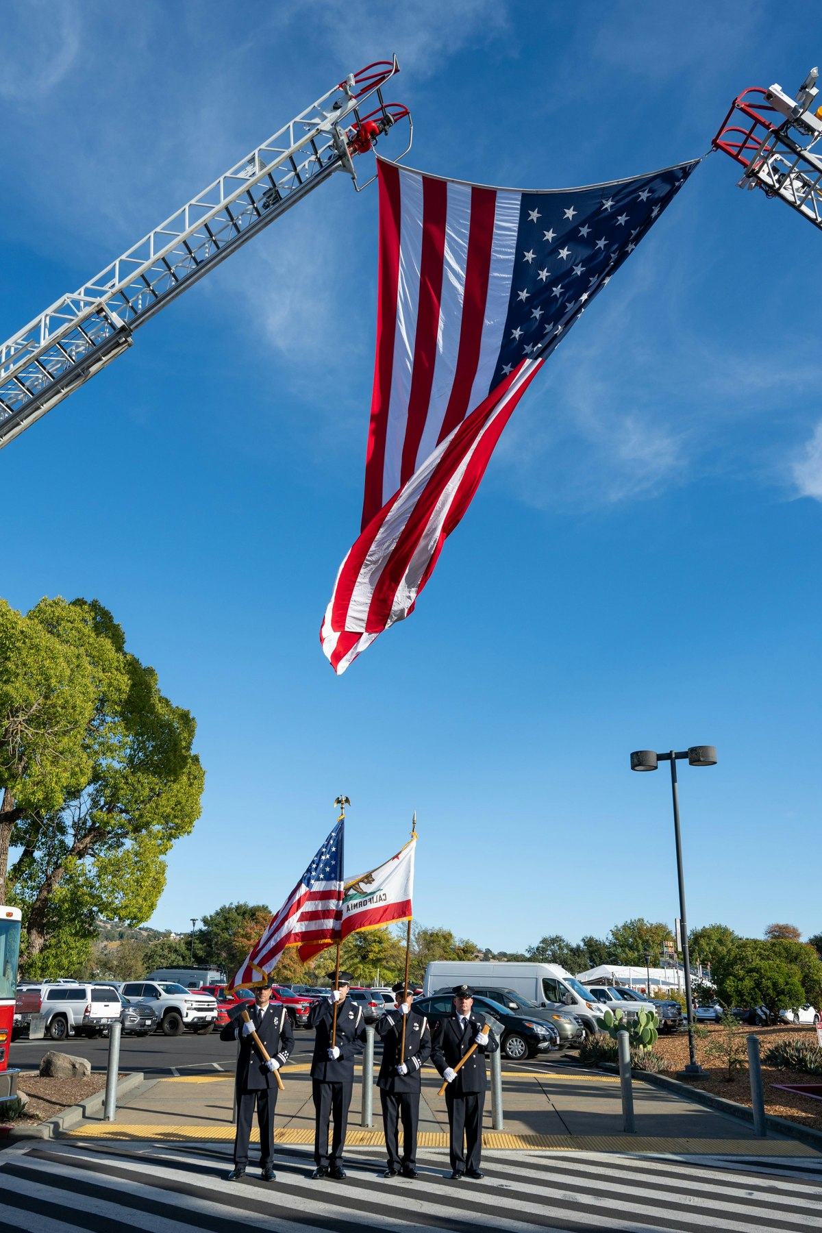 A large American flag hangs between fire truck ladders; uniformed individuals stand below, holding U.S. and California flags on a sunny day.