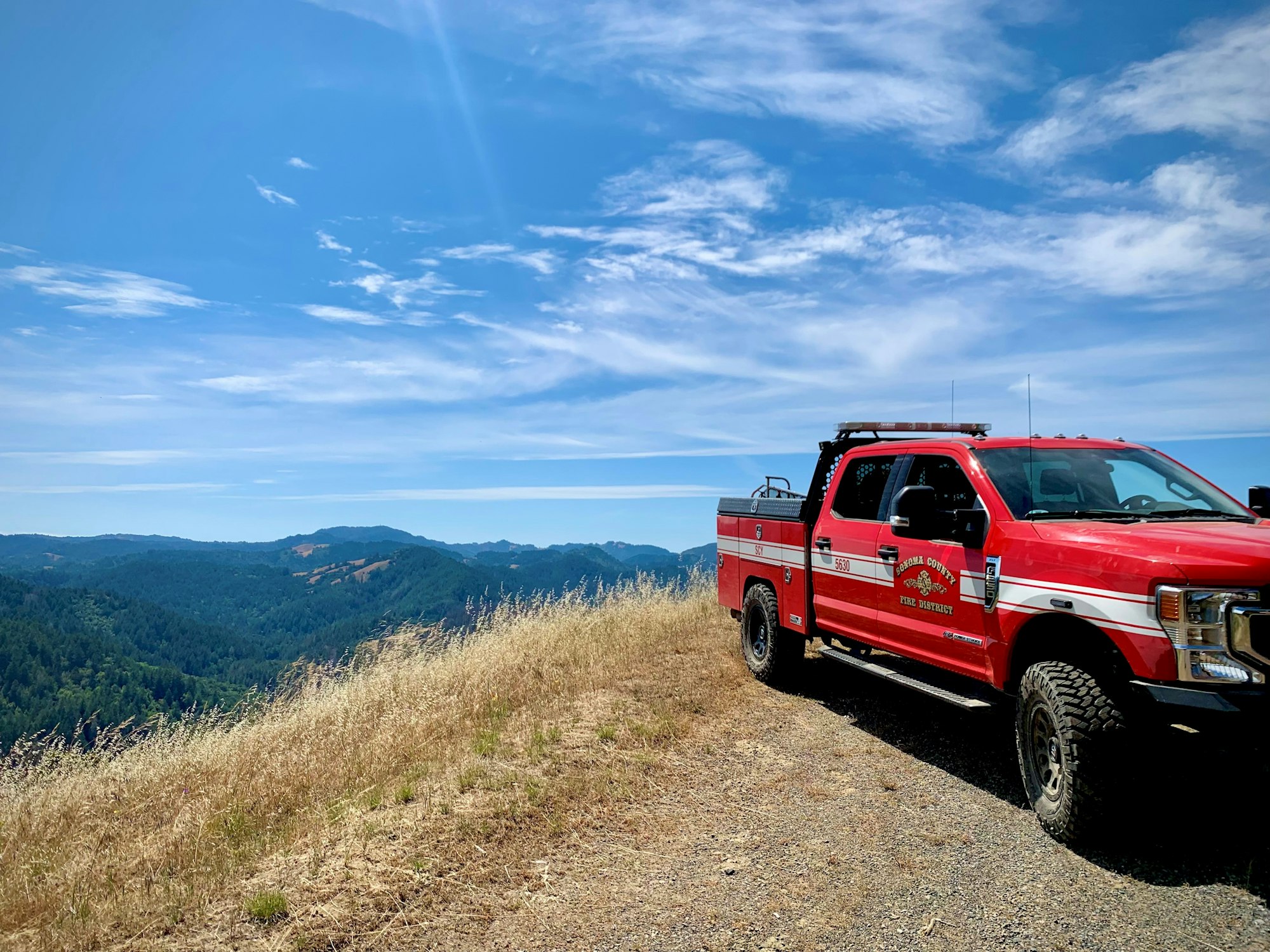 Red fire truck parked on a hilltop with a scenic view of rolling green hills and a bright blue sky.
