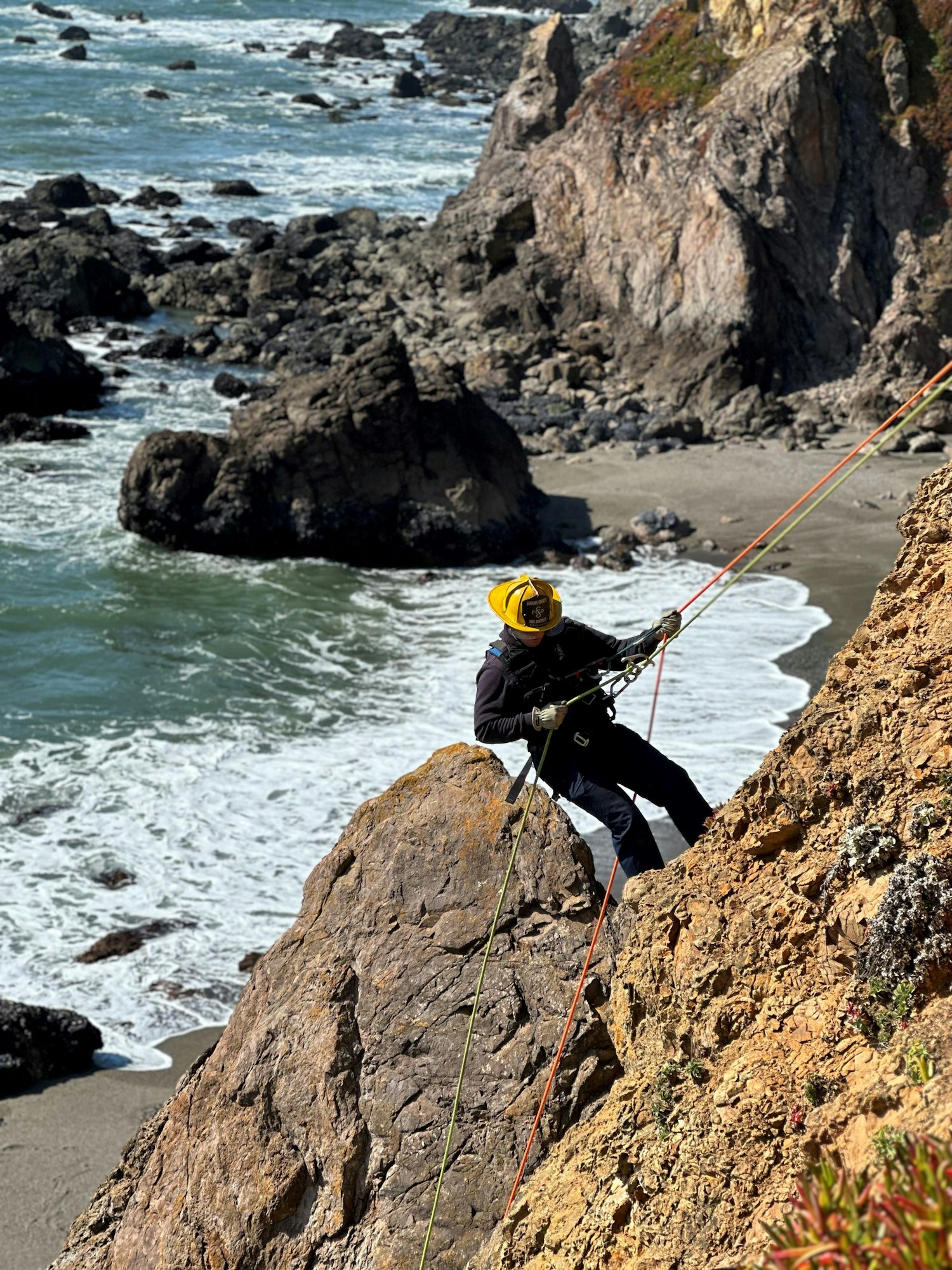 A person in a yellow helmet rock climbing by the ocean with ropes, surrounded by rocky cliffs and waves.