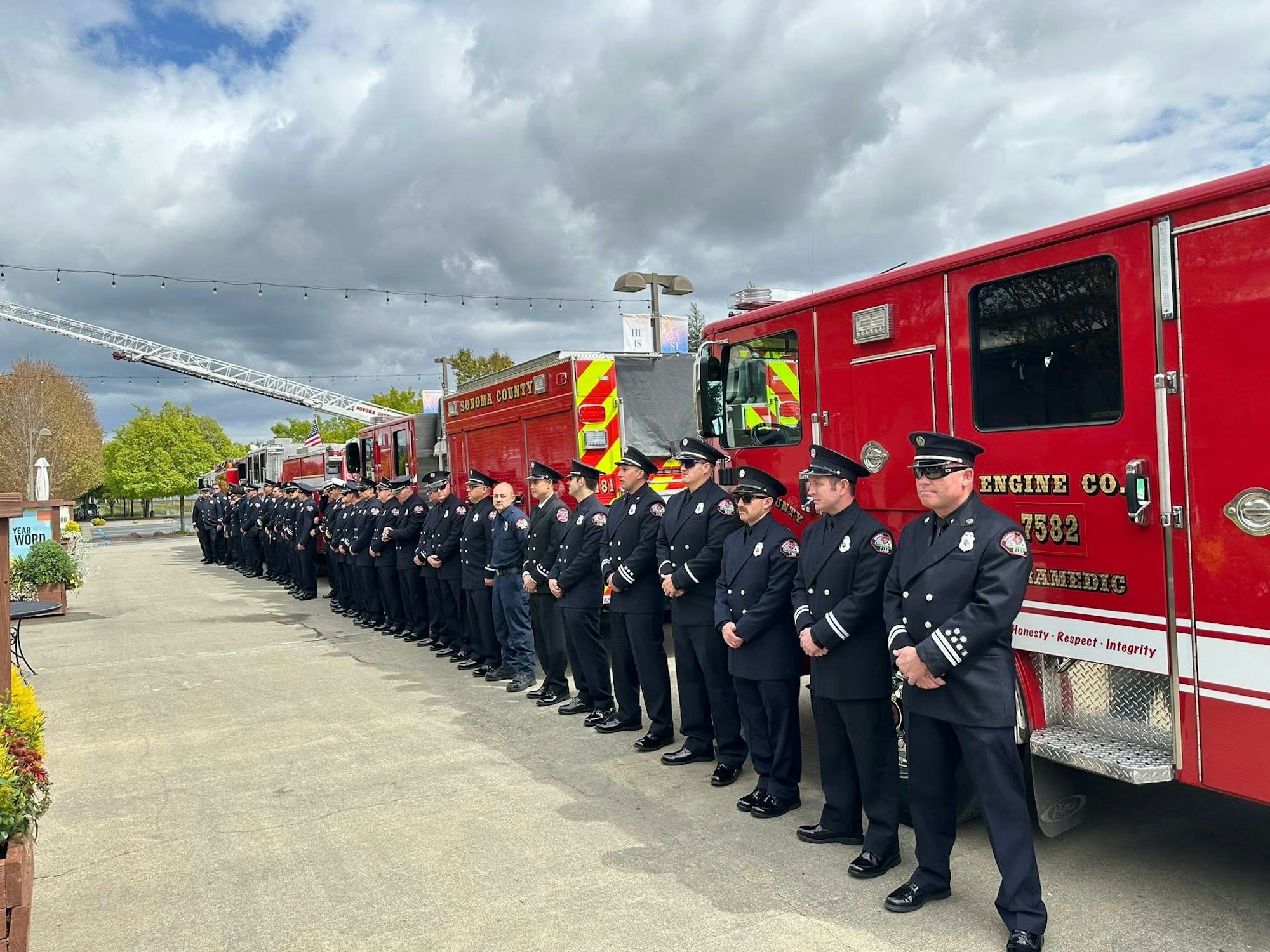 A line of firefighters in uniform stand beside red fire trucks under a cloudy sky.