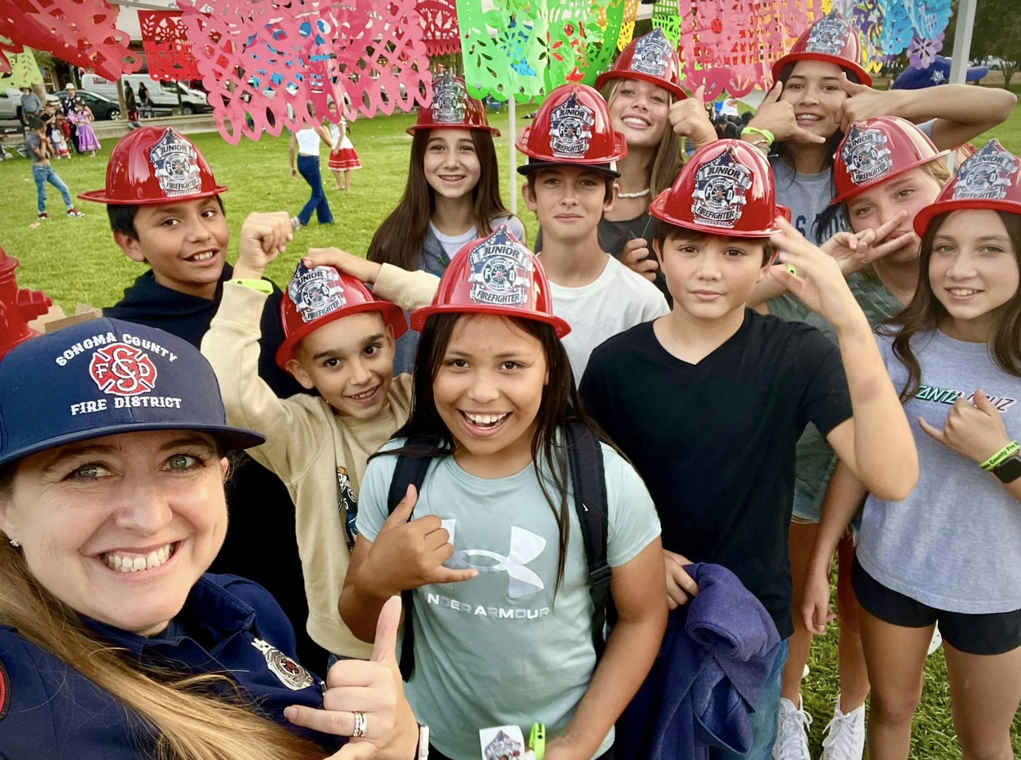 A group of kids wearing red firefighter helmets, posing with a person in a fire service cap under colorful decorations.