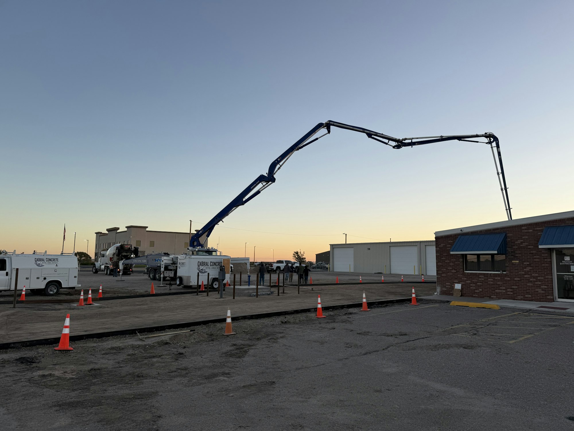 Construction site with concrete pump trucks, traffic cones, and buildings at sunset.
