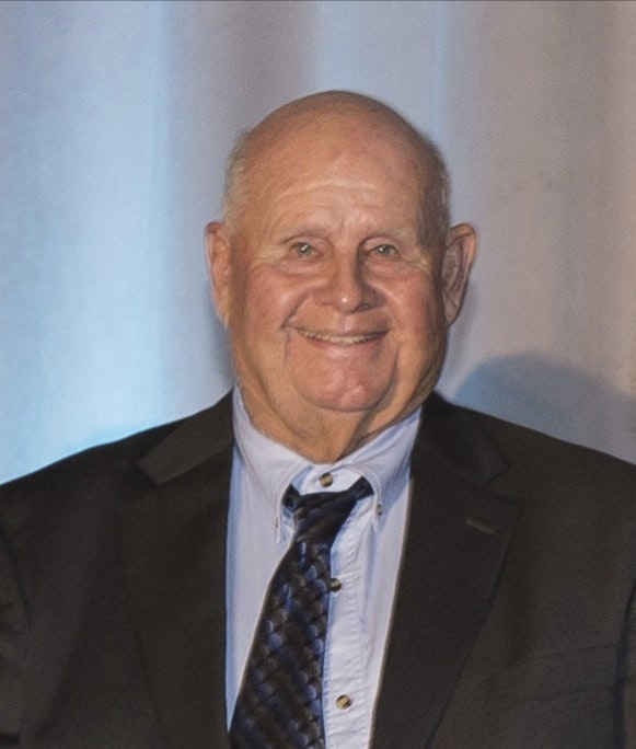 The image shows an older man in a suit smiling warmly against a softly lit background.