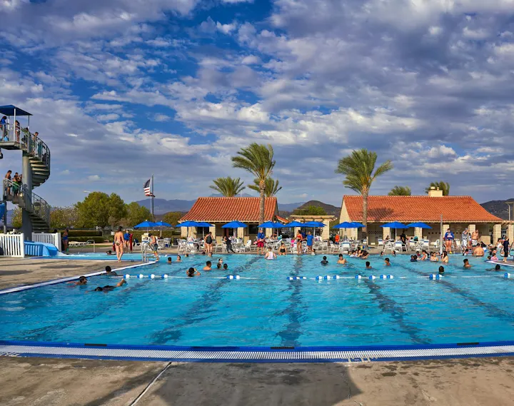 A sunny pool scene with people swimming, a water slide, and palm trees, surrounded by lounge chairs and blue umbrellas.