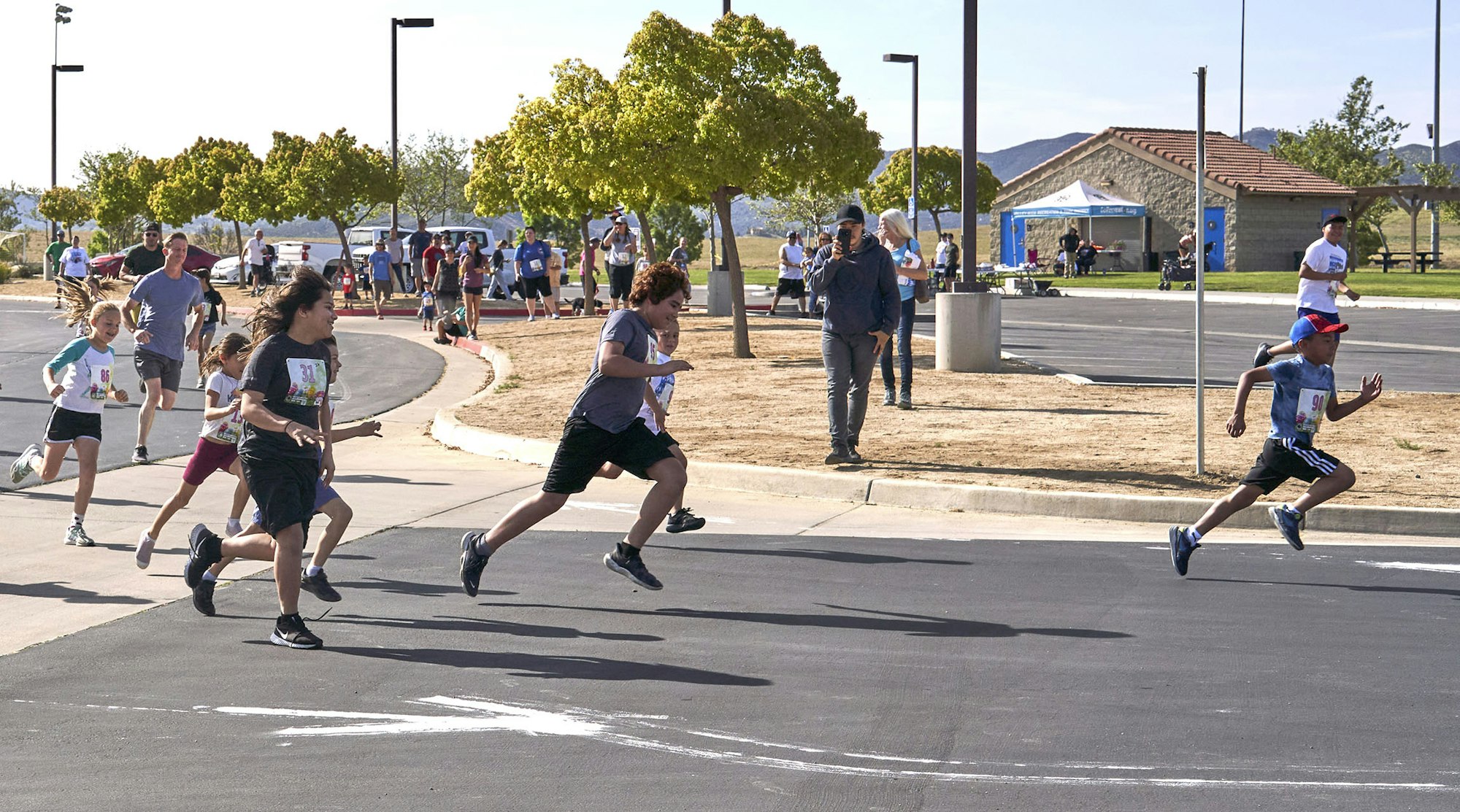 A group of kids and adults participating in a running event at a park, with some sprinting and others walking in the background.