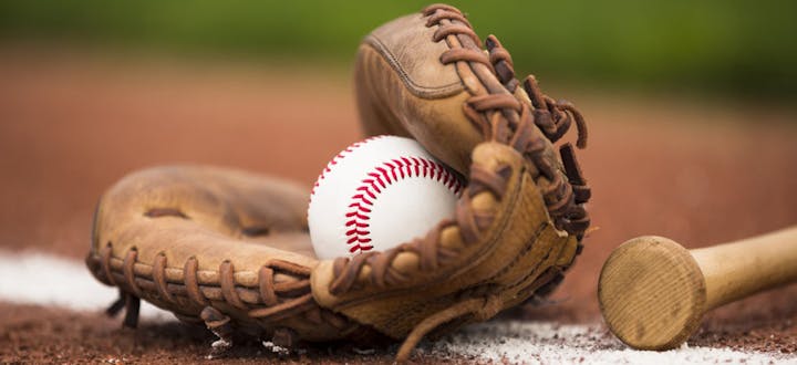 A baseball resting in a glove with a wooden bat nearby, set on a dirt field, suggesting a scene from a baseball game.