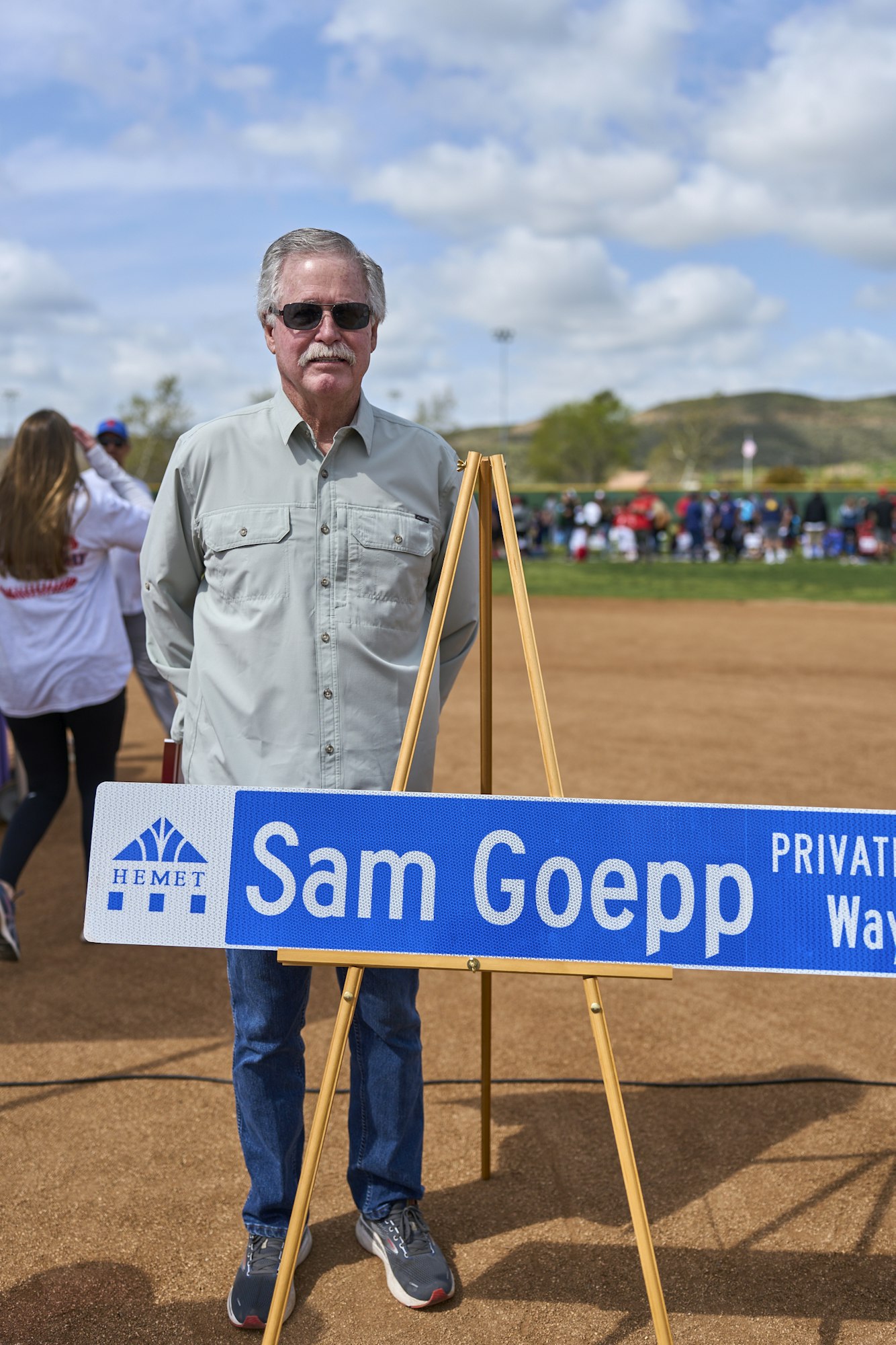 A man in sunglasses stands behind a sign on an easel that reads "Sam Goepp." People and a field are in the background.