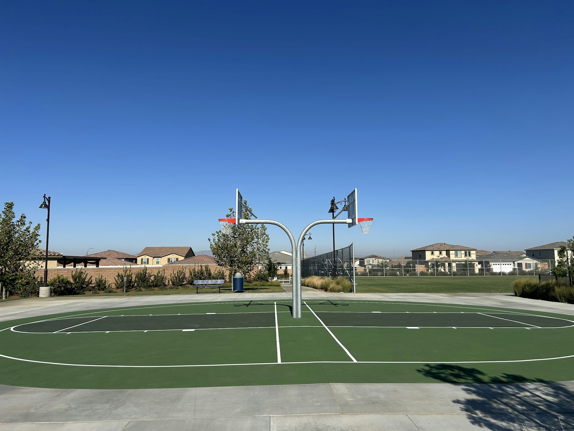 An empty basketball court with two hoops under a clear blue sky, surrounded by houses and greenery.