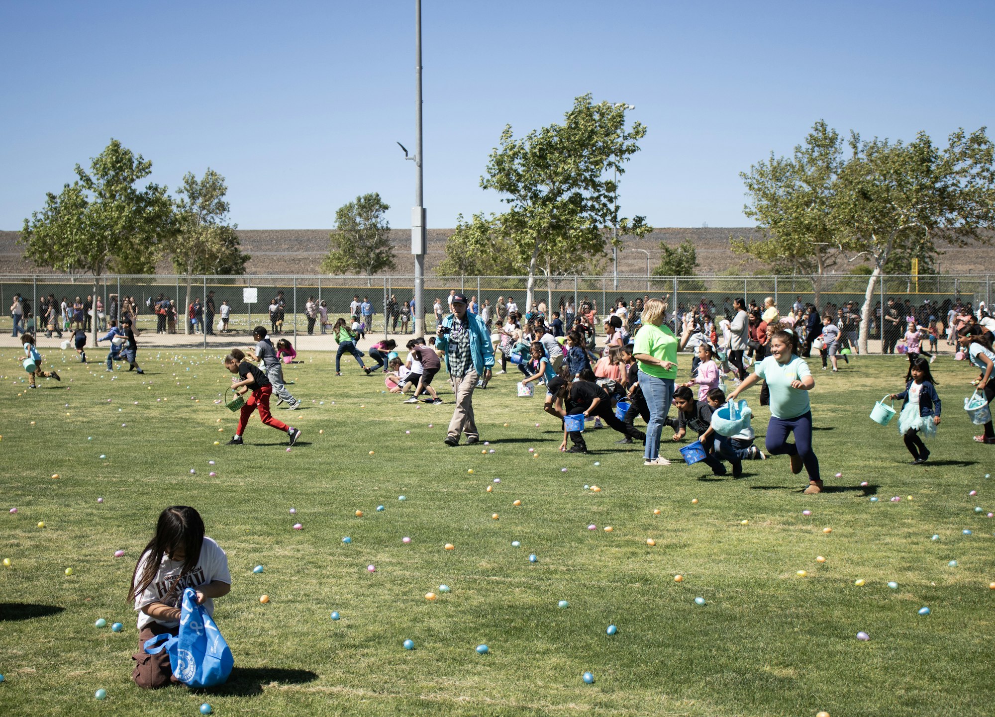 Children are participating in an egg hunt in a park, collecting colorful eggs while adults watch from the sidelines.