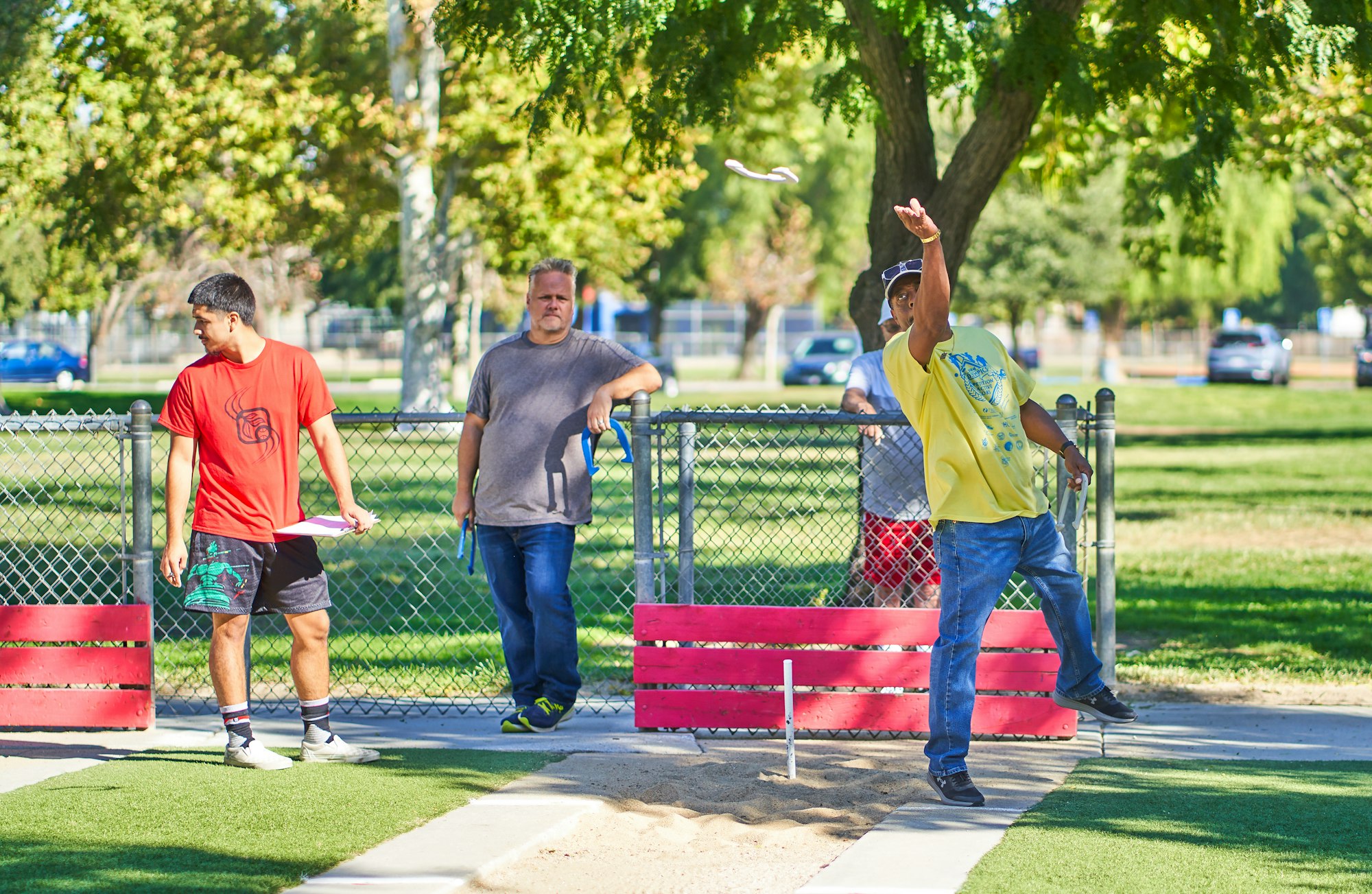 Three men playing disc golf in a sunny park, with one disc in mid-flight.
