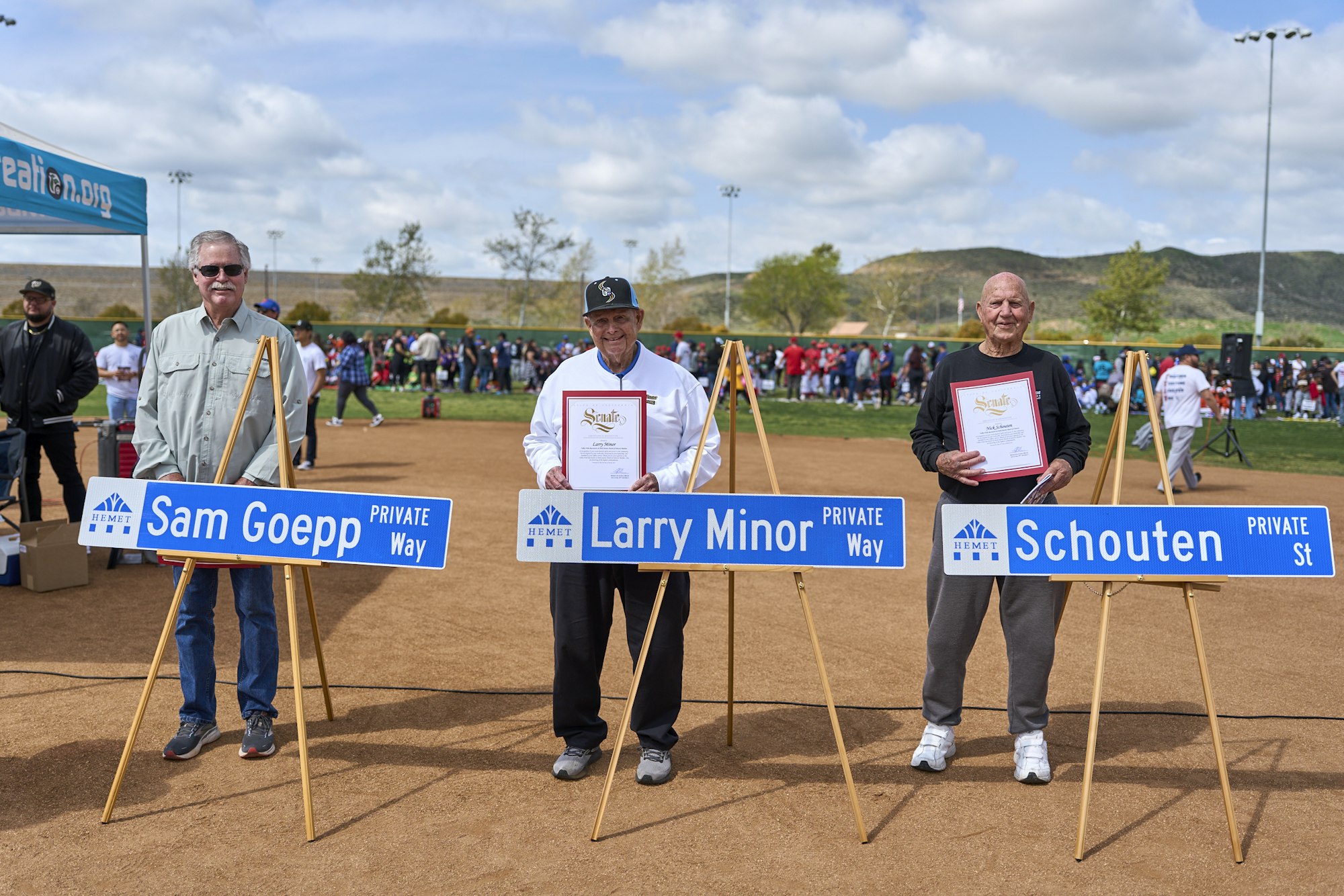 Three men stand holding certificates behind street signs with their names at an outdoor event.