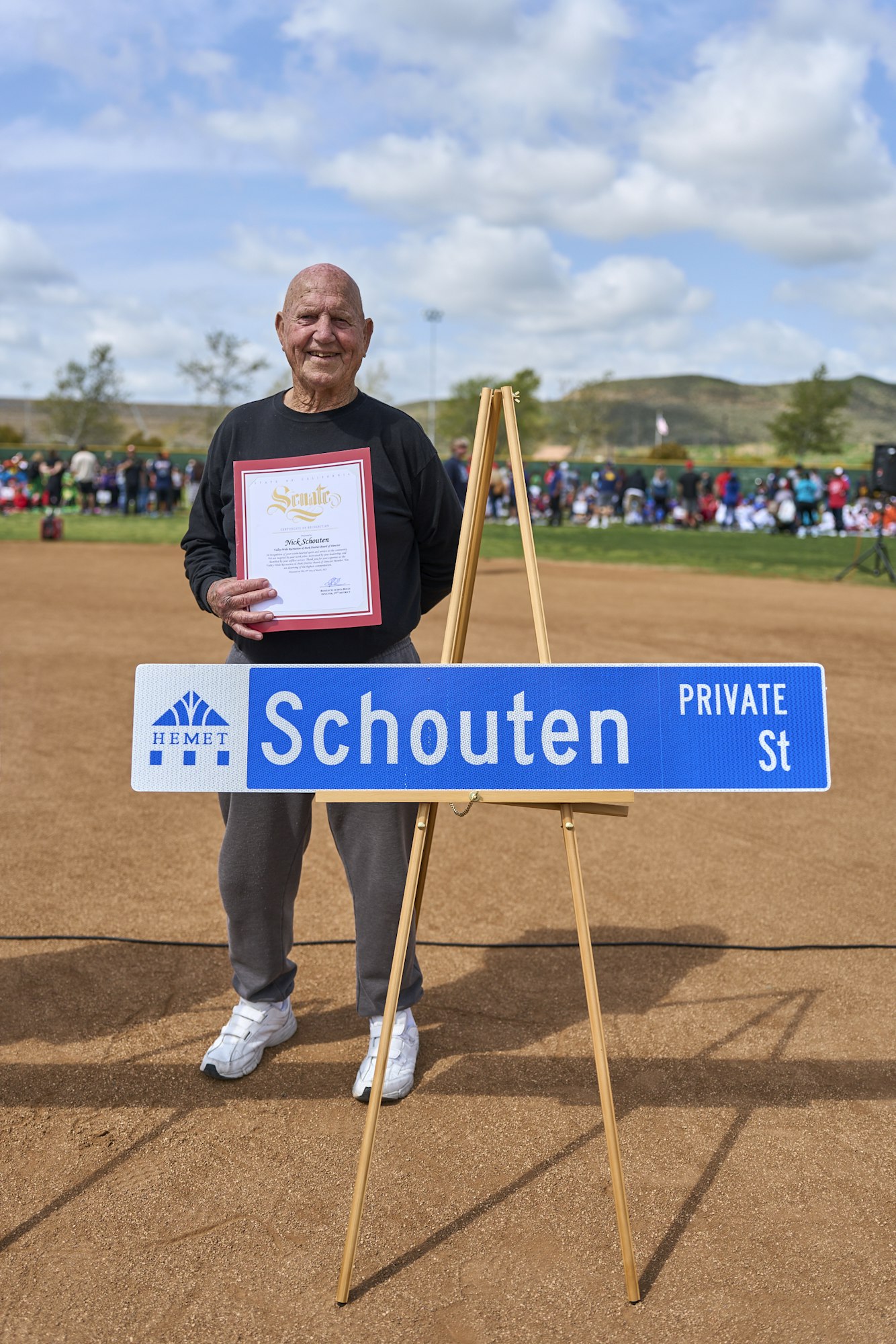 A person holding a certificate, standing by a sign that reads "Schouten Private St," with a crowd and scenic background.
