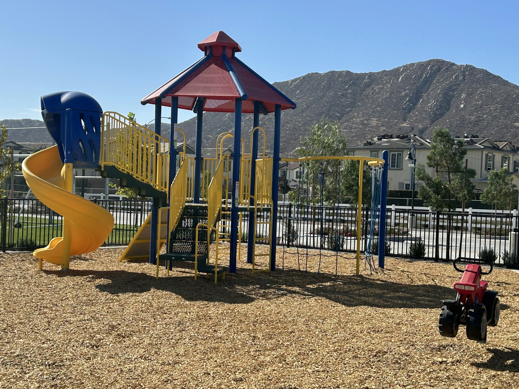 The image shows a colorful playground with a slide, climbing structure, swings, and a toy tractor in a sunny setting.
