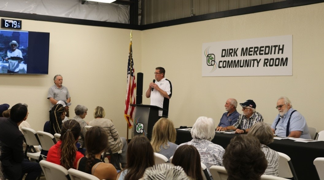 A man speaks at a podium in the "Dirk Meredith Community Room" to an audience, with seated panelists and an American flag nearby.