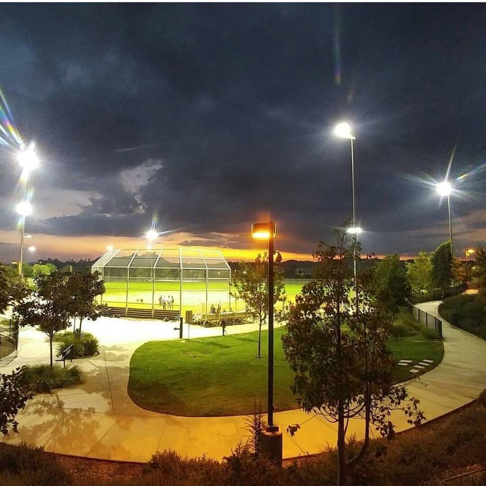 A lit baseball field under a dramatic sky, with dark clouds and a sunset, surrounded by trees and pathways.