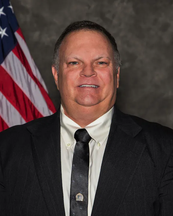 A man in a suit and tie smiles for the camera, with an American flag in the background.