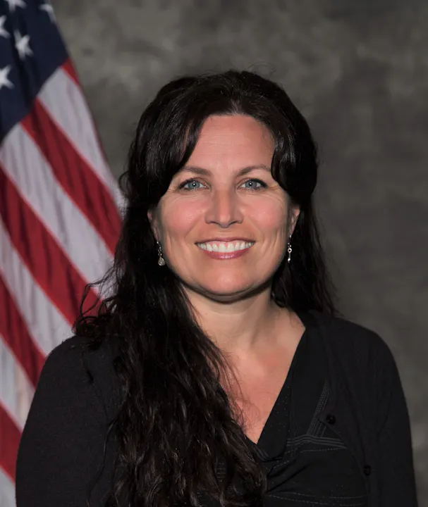 A woman with long dark hair smiles in front of an American flag backdrop, wearing a black top and earrings.